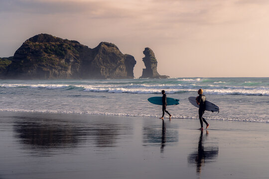Two surfers in wetsuits walk along the beach with their surfboards after a surf session in Piha, Auckland, New Zealand, with Lion Rock in the background.