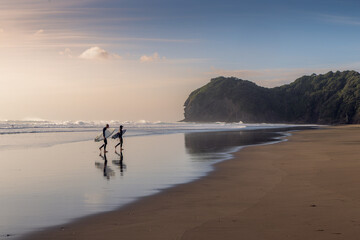 Two surfers walk along the beach with their boards at Piha Beach in Auckland, New Zealand. They are...