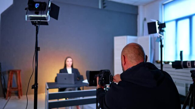 Rear view of a cameraman working in modern studio. Soffits light the woman sitting at desk in blurred backdrop.