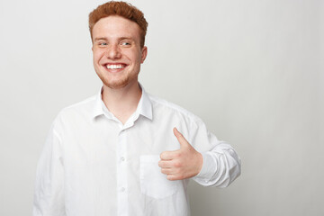 Smiling young man with red hair and beard wearing white shirt shows thumbs up gesture. Positive...