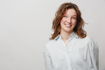 Obraz premium Smiling young woman with curly brown hair wearing white shirt looks up with cheerful expression. Studio portrait on light background. Concept of happiness, positivity, and casual lifestyle.