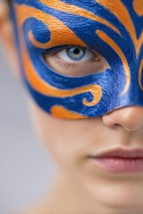 Obraz premium Young woman wearing a blue and orange decorative mask, showcasing intricate designs, with a focus on her expressive eye and smooth skin in a neutral background setting