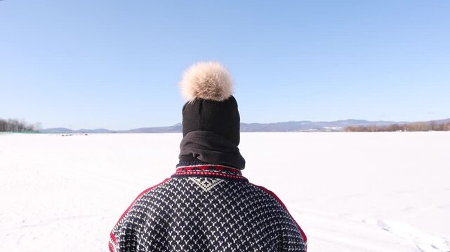 Mature woman dressed warmly wearing sweater and toque with pom pom outdoors in winter looking out over frozen snow covered prairie leading to mountains with ski slopes apparent.