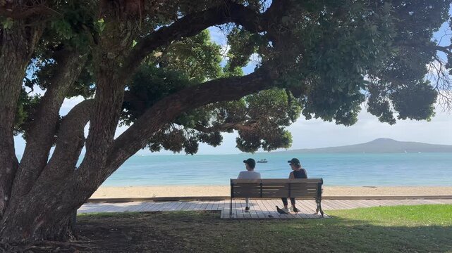 Two people sit on a bench at St Heliers, enjoying the view of Rangitoto Island on a sunny day.