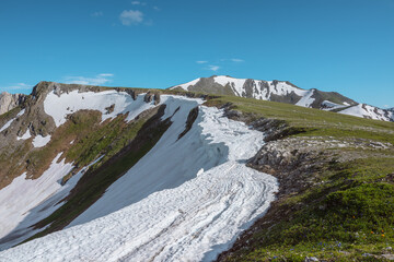 Scenic alpine sunlit view along green grassy ridge with white snow cornice and rocky sheer crags in bright sun under blue sky. Awesome landscape with sharp rocks and snowy mountain range in sunny day. © Daniil
