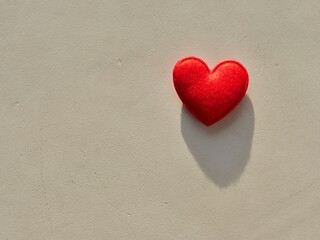 Fototapeta premium Red felt heart on a neutral stone background with sunlight and long shadow. Minimalist romantic concept with a handmade fuzzy wool heart. Top view of a red fiber heart for Valentine's Day, wedding,