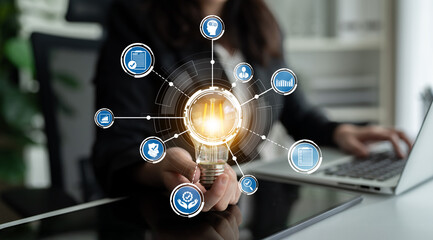 A businesswoman presents a light bulb that represents innovation, surrounded by digital icons. This image conveys creativity and modern technology in a corporate environment. Crux © InfiniteFlow