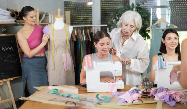 Older female instructor standing beside young woman stitching at machine, offering help during group sewing practical class in cozy professional workshop..