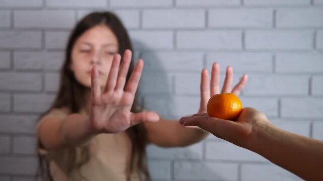 Teenager refusing to eat a tangerine. Dissatisfied young girl showing a stop gesture with her hands and refusing to accept a tangerine fruit, demonstrating a concept of food allergy, dislike