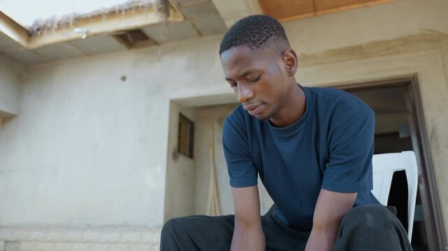 Wide shot black man after chores, seated on house steps, concrete stoop and unfinished plaster exterior, navy shirt, downward gaze, hands resting on knees, midday light, quiet pause between tasks,