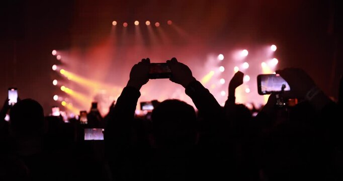 Audience members in silhouette use their phones to record a concert. Intense orange and red stage lighting creates a high-energy atmosphere.