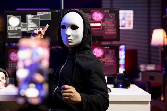 Hacker hidden in neon lit hideout films video ultimatum with smartphone, sending it to foreign governments. Masked evil man threatens retaliation against foreign military targets