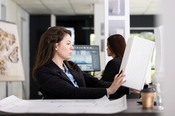 Caucasian architectural designer examines scaled 3D building prototype at desk, crosschecking...