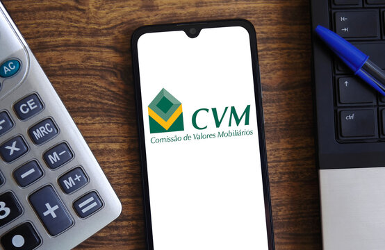 The CVM (Brazilian Securities and Exchange Commission) logo is displayed on a smartphone screen with a laptop and calculator in the background. Concept of finance, economics, and investment.