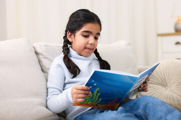 Fototapeta premium Cute little girl reading book on couch at home