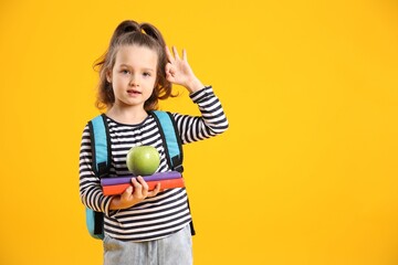 Fototapeta premium Cute schoolgirl with backpack, books and apple showing OK gesture on yellow background. Space for text