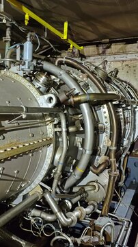 Closeup turbine assembly with curved pipes and wiring, industrial hangar maintenance scene showing coils, flanges, bolts, insulated tubing and yellow support bracket, detailed metal surfaces and worn