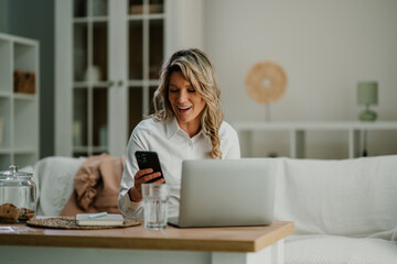 Woman working remotely at home, smiling and communicating using smartphone and laptop for business...