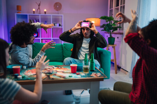 Diverse friends having fun playing charades game at home party, using mobile phone for guessing word