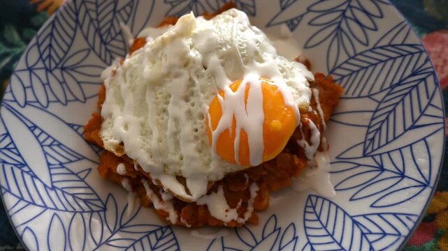 A close-up shot of red chilaquiles topped with a fried egg and Mexican crema, served on a patterned ceramic plate.