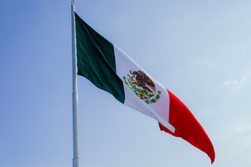 National flag of Mexico featuring the eagle coat of arms waving in the wind against a clear blue...