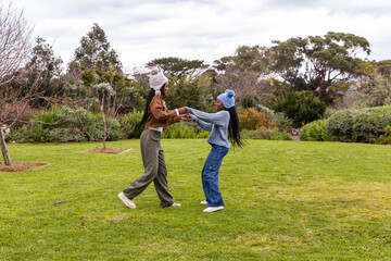Two teenage African American females holding hands, spinning on lawn at park with beanies, sneakers