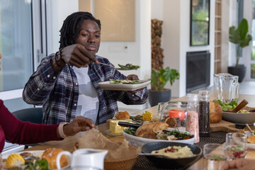 Naklejka premium African American man with dreads in plaid serving salad with spoon at family meal at home