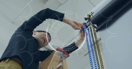 Asian in black top, khakis adjusting round ceiling device on ladder in workshop, with pulley straps