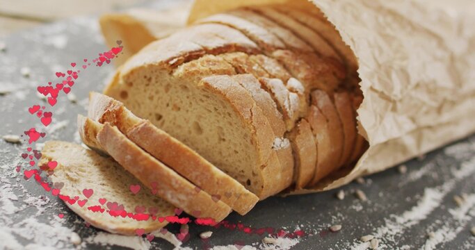 Showing rustic sliced loaf resting in crumpled paper bag on slate, flour-dusted with pink hearts