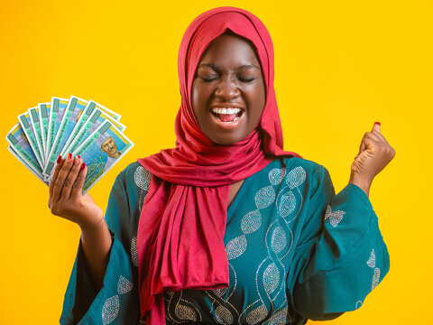 A joyful Black Muslim woman in a teal patterned dress and red hijab celebrates while holding a fan of Djibouti Francs. Set against a bold yellow background, she expresses pure excitement and success.