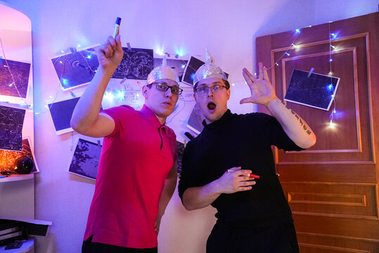 Two Men Wearing Foil Hats In Study Gesturing At Star Maps And Glowing Lights, Playful Alarm And Surprised Expressions, Amateur Ufologists Preparing Theory Session With Posters And String Lights