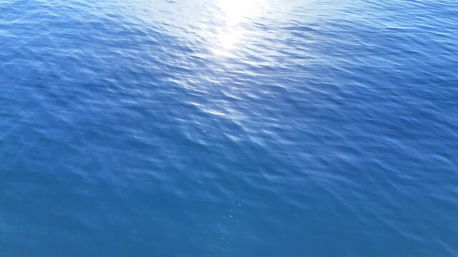 Cinematic drone flight over calm ocean water with bright sun glare and soft rolling ripples, minimal blue marine landscape background in serene daylight
