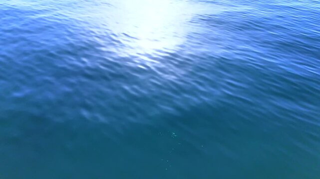 Cinematic drone flight over calm ocean water with bright sun glare and soft rolling ripples, minimal blue marine landscape background in serene daylight
