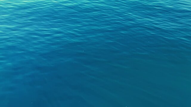 Top view drone footage gliding above deep blue sea with sparkling sun reflection and soft wave patterns, peaceful ocean background in bright natural light