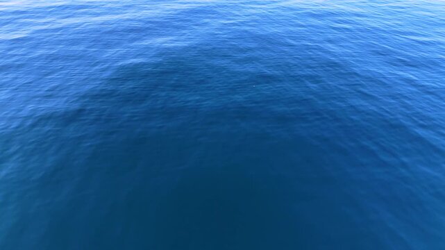 Aerial drone shot flying over calm blue ocean surface with shimmering sunlight reflection and gentle ripples, cinematic seascape background in natural daylight