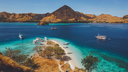 Aerial view of a tropical island with turquoise waters, white sand, anchored boats, and golden hills under a clear blue sky. © Lylys