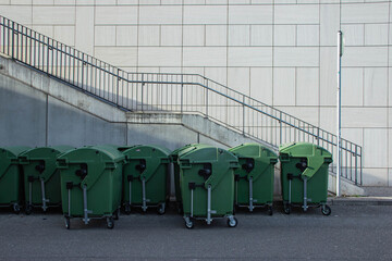 Row of green waste bins on wheels positioned against a concrete wall with a staircase and railing in the background, urban setting with clear lines and textures