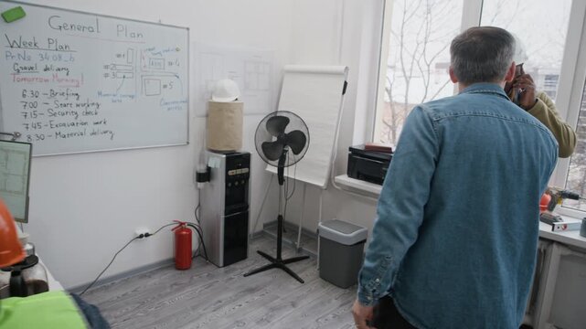 Tracking shot of middle-aged Caucasian construction worker entering office, pouring water from cooler into glass, talking to colleagues and putting on hardhat and safety vest before work