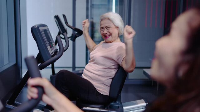 Happy senior Asian woman exercising on recumbent bike at gym, smiling and cheering with trainer. Healthy lifestyle, active aging, and fitness for elderly people in modern sports club facility.