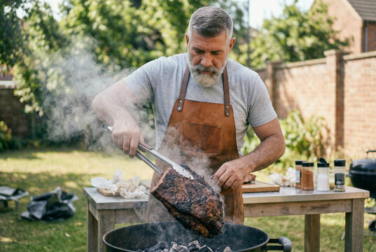 Rugged pitmaster lifting a large piece of smoked meat from a smoky outdoor barbecue grill.
