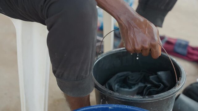 Black hands washing clothes in bucket, outdoor porch scene, wringing dark garment with soapy water, seated on plastic chair, patient rhythm, home caretaker gesture, tactile detail, subtle sunlight