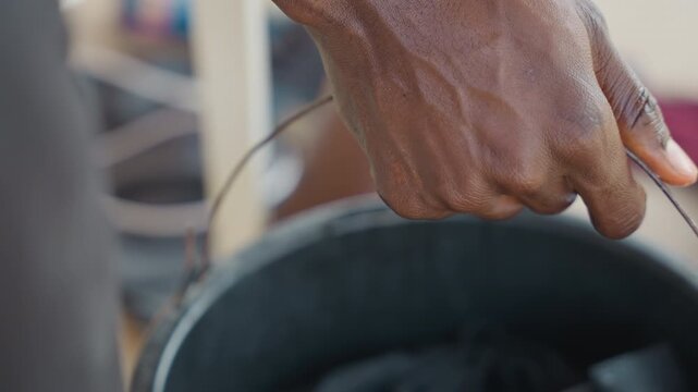 Black hand holding bucket handle closeup, soaked fingers gripping metal rim, dark laundry in soapy water, courtyard or porch setting, callused skin and steady pressure, laundry worker motion,