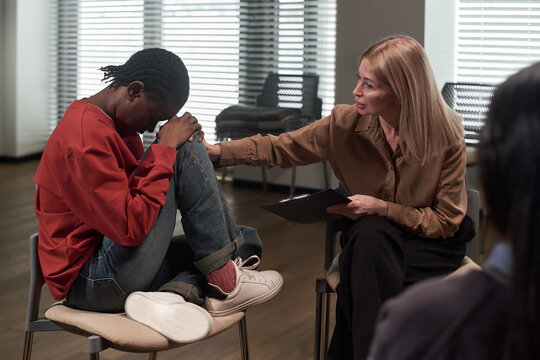 Caucasian middle aged woman comforting Black teenager sitting with knees up and head down during counseling session, therapist gently touching her arm, emotional support concept