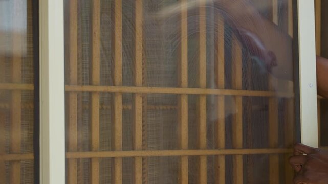 Homeowner cleaning window screen closeup soapy sponge wipes mesh and wooden frame, sunlight filtering through porch, methodical wiping motion, seasonal house chores, calm domestic routine, textured