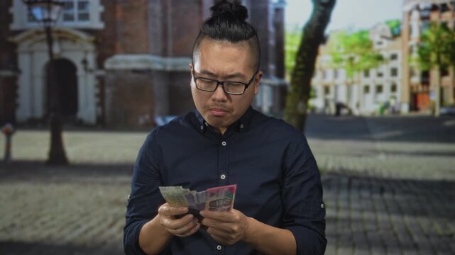 Man counting indonesian rupiah banknotes on a cobbled city street, frowning while holding cash and checking notes; financial worry.