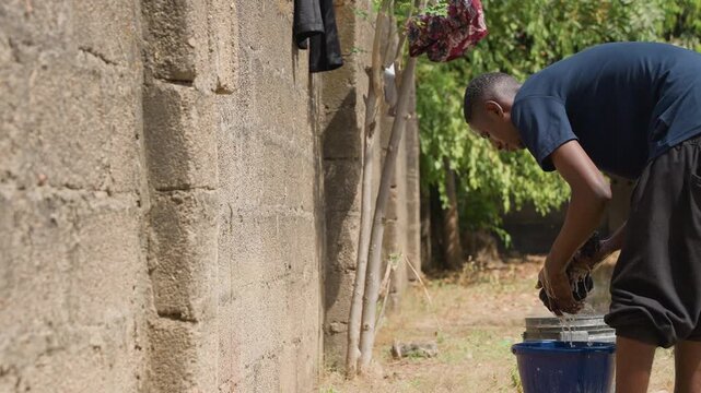 Man washing black clothes in blue bucket beside house, bending and wringing garments, sudsy water and drops on concrete, focused posture, sunlit backyard, manual routine and practical household chore
