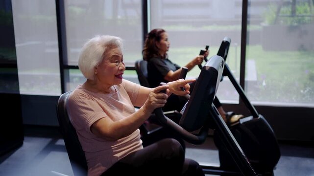 Two happy senior Asian women exercising on recumbent stationary bikes at gym, smiling and pointing at screen. Concept of active aging, healthy lifestyle, and fitness for elderly people in center.