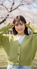 満開の桜の下で春らしいグリーンのカーディガンをまとい、笑顔でポーズをとる女性