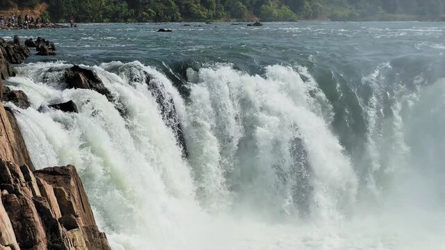 closeup shot of Dhuandhar Waterfall at Bhedaghat, Jabalpur, as the Narmada River plunges over rocky cliffs, with visitors visible in the distance near the river edge.