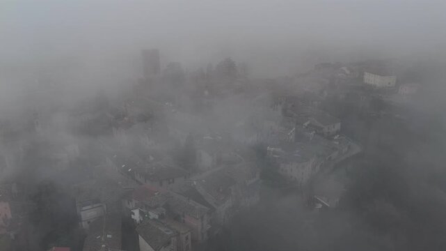 Castell&rsquo;Arquato historic center shrouded in dense fog, revealing medieval tower, church and stone building through mist over hillside landscape in Emilia-Romagna, Italy, drone slow reveal shot.